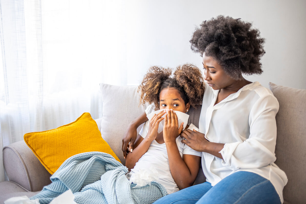 Mother comforting her sick child on the couch as the child uses a tissue—symptoms could be from allergies or a cold.